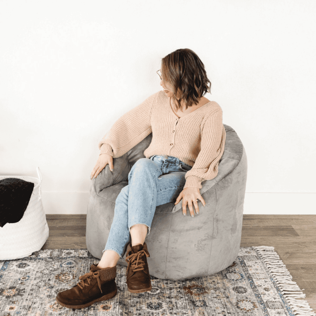 A young woman with short hair is seated in a light grey bean bag chair, wearing a beige knitted sweater and light blue jeans. She appears relaxed, with a cosy interior featuring a patterned rug and a woven basket in the background.