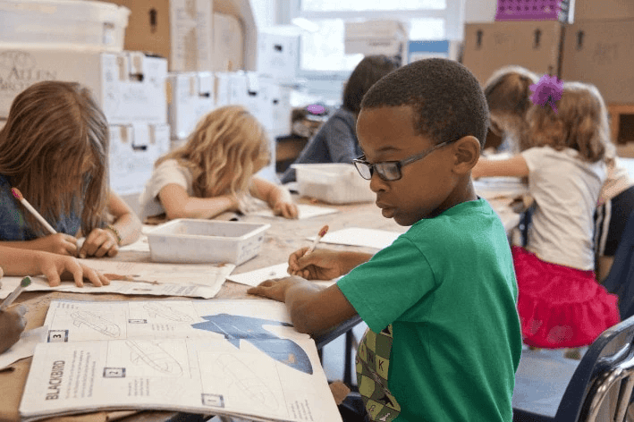 A group of elementary-aged children, including a boy in glasses and a teal shirt, are seated around a table working on papers. The table holds worksheets, plastic trays, and other materials, with stacked cardboard boxes suggesting an inclusive classroom setting.