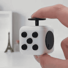 A hand fiddles with a fidget cube, drawing focus against the blurred background of a white wall, which includes a small Eiffel Tower model and a tiny bottle. The blurred backdrop enhances attention on the fidget cube in use.