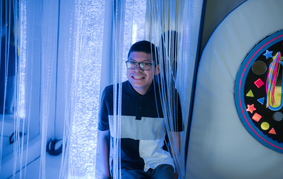 A young man with dark hair and glasses smiles warmly in a sensory room surrounded by soft, light-blue fibre optic curtains. The calming space contrasts with vibrant, multicoloured geometric wall designs, blending soothing light with stimulating sensory elements.