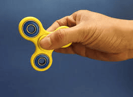 A hand holds a bright yellow fidget spinner, ready to spin. The spinner’s three arms, each capped with a blue ring, stand out against the cool grey background, highlighting its vibrant colours and creating a sense of calm and anticipation.
