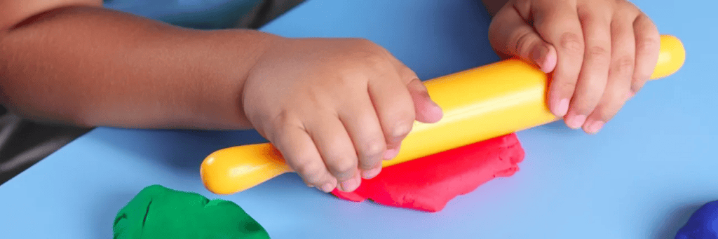 The image shows a child’s hands rolling out bright red playdough using a yellow rolling pin on a blue surface. Green playdough is also present nearby, suggesting a playful and creative activity involving tactile materials, ideal for developing fine motor skills.