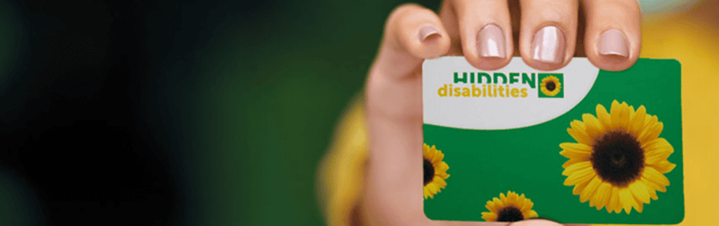 The image shows a close-up of a hand holding a green card featuring two sunflowers and the words "Hidden Disabilities" written in white and yellow text. The sunflower imagery and green background are symbolic of the Hidden Disabilities Sunflower scheme.